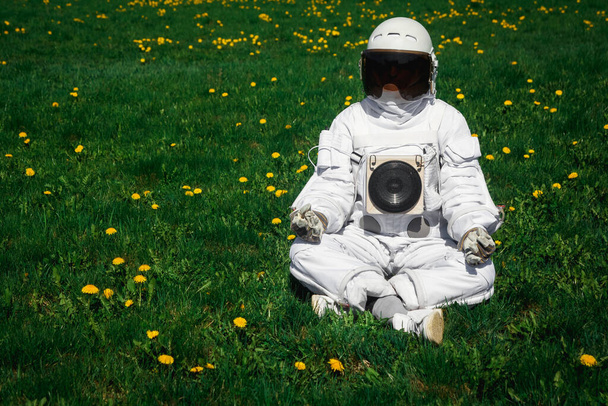 ヘルメットの未来的な宇宙飛行士は瞑想的な位置の間で緑の芝生の上に座っている. - 写真・画像