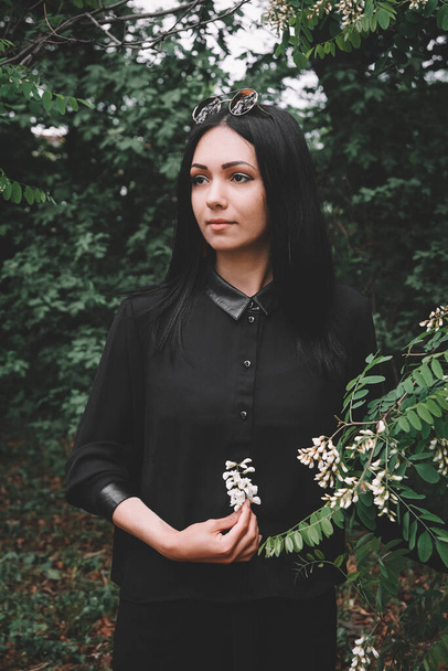 Beautiful girl in black clothes and glasses from the sun holds a white flower in her hand against the background of green leaves - Fotó, kép