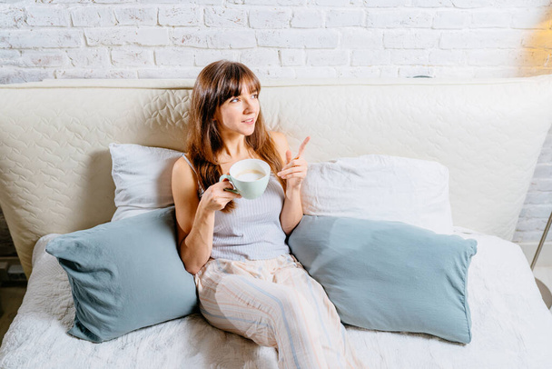 白い寝具と枕の中に座っている間、コーヒーを飲みながらパジャマを着た魅力的な若いブルネットの女性。朝、レジャー、冬と人々の概念 - 写真・画像