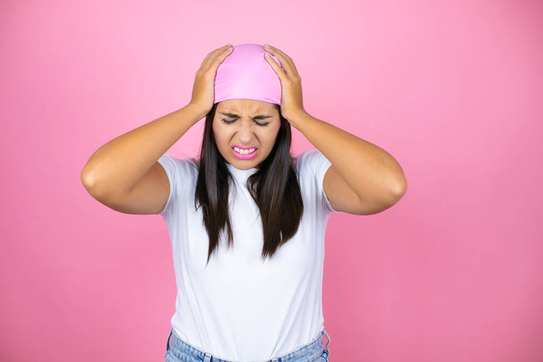 Молодая красивая женщина в розовом платке на изолированном розовом фоне страдает от головной боли отчаянно и стресс, потому что боль и мигрень с руками на голове - Фото, изображение