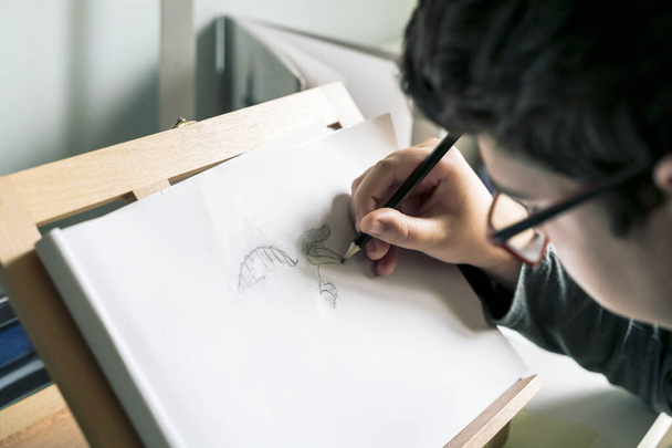 concentrated teenager draws a dinosaur in pencil in his sketchbook on a wooden lectern. - Photo, Image