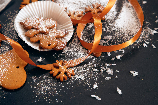 Christmas freshly baked gingerbread cookies sprinkled with powdered sugar with snowflakes and ribbon on black background. Holiday food, homemade baking, Christmas and New Year traditions. - Photo, Image