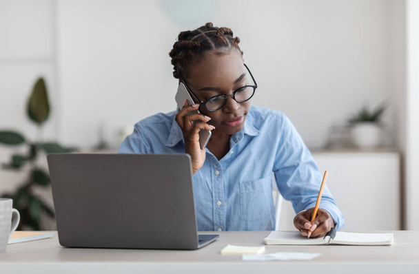 Czarna kobieta pracownik biura rozmawia przez telefon komórkowy z klientem i robi notatki - Zdjęcie, obraz