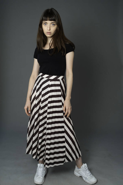 Studio portrait of a pretty brunette woman in a black t-shirt and striped skirt, standing against a plain grey background, looking at camera - Foto, Imagem