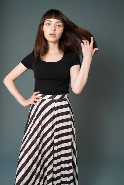 Studio portrait of a pretty brunette woman in a black t-shirt and striped skirt, standing against a plain grey background, looking at camera - Foto, imagen