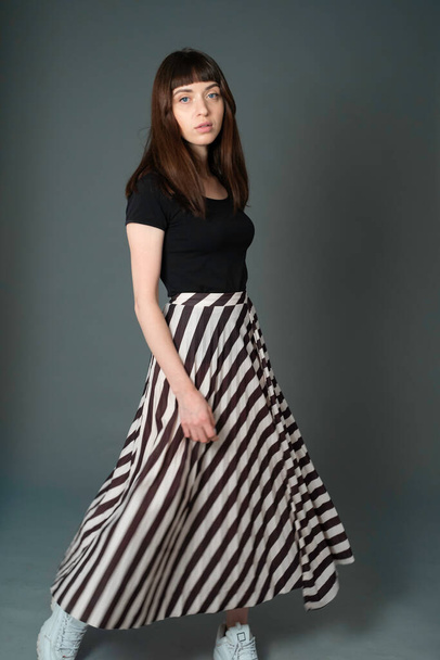 Studio portrait of a pretty brunette woman in a black t-shirt and striped skirt, standing against a plain grey background, looking at camera - Fotó, kép