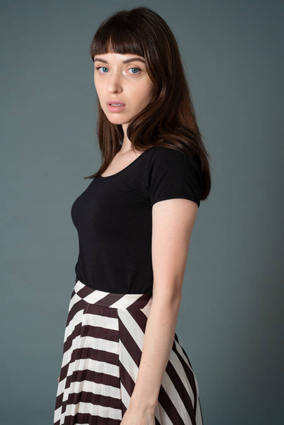 Studio portrait of a pretty brunette woman in a black t-shirt and striped skirt, standing against a plain grey background, looking to the side - Foto, imagen
