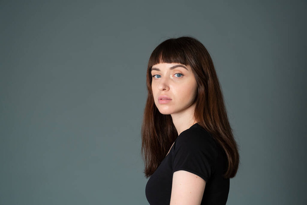 Studioporträt einer hübschen brünetten Frau in schwarzem T-Shirt, die halb gedreht vor einem schlichten grauen Hintergrund steht und in die Kamera blickt - Foto, Bild