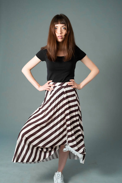 Studio portrait of a pretty brunette woman in a black t-shirt and striped skirt, standing on one foot, against a plain grey background, looking at the camera - Zdjęcie, obraz
