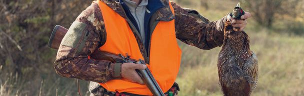 A man with a gun in his hands and an orange vest on a pheasant hunt in a wooded area in cloudy weather. A hunter with a pheasant in his hands. - Photo, Image