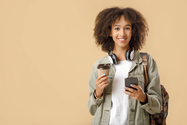 Der moderne studentische Hipster macht nach Vorlesungen Kaffeepause. Fröhliche Afroamerikanerin mit kabellosen Kopfhörern, Rucksack und Tasse Kaffee zum Mitnehmen tippt auf dem Smartphone - Foto, Bild