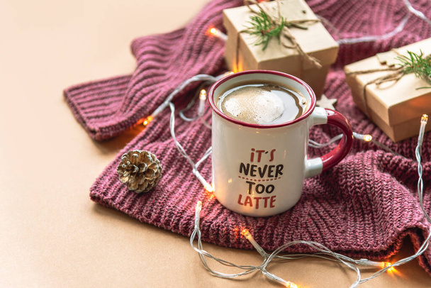 Egy csésze kávé habbal és karácsonyi ajándékokkal lila kötött pulóveren bézs alapon, koszorúval. Közelről. - Fotó, kép