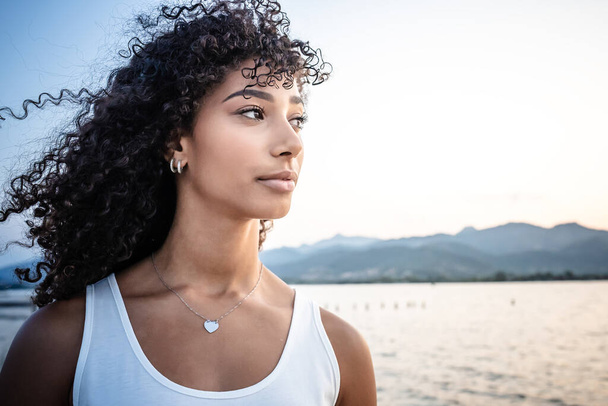 Beautiful Hispanic young woman head and shoulders portrait with long curly dark hair in white undershirt looks to the sun at dusk with lake and mountains background - Human beauty and that of nature - Fotografie, Obrázek