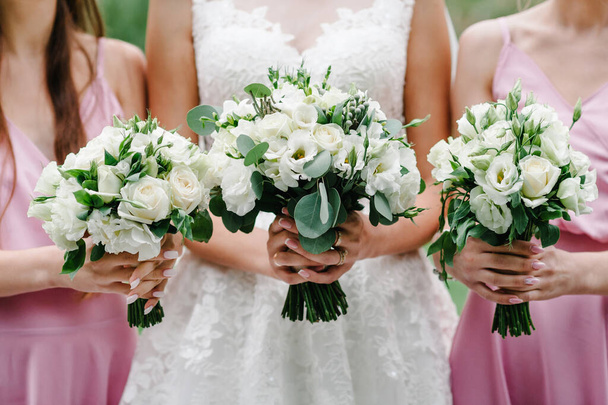 Nevěsta a družičky v elegantních pastelově růžových šatech stojí a drží v přírodě kytice květin a zeleně. Mladé krásné dívky drží svatební kytice venku. - Fotografie, Obrázek