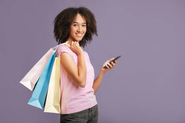 Moderni ostoksia, alennuksia ja tarjouksia. Hymyilevä afrikkalainen amerikkalainen nainen värillinen paperi pussit katsot kameraan, tilalla älypuhelin ja saa cashback - Valokuva, kuva