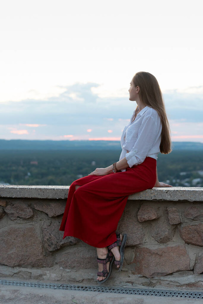 Young woman in red skirt sits on observation deck, nature on background. Vertical frame. - Photo, Image