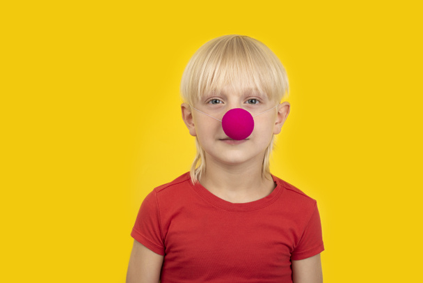 Портрет светловолосого мальчика с клоунским носом в красной рубашке на жёлтом фоне - Фото, изображение