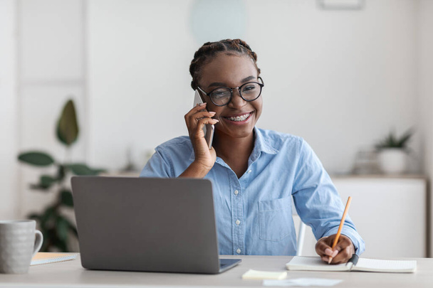 Estagiário de escritório. Mulher negra conversando no celular e tomando notas no local de trabalho - Foto, Imagem