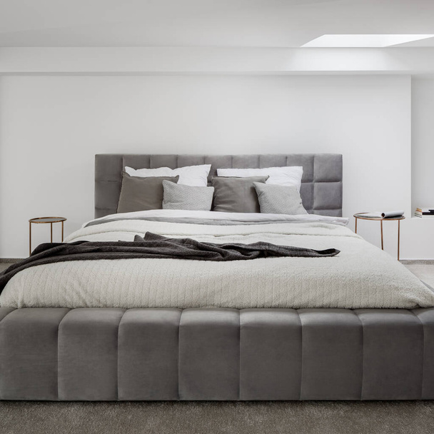 Большая кровать с серой и стеганой рамой в спальне с белыми стенами, серым полом и окном потолка - Фото, изображение