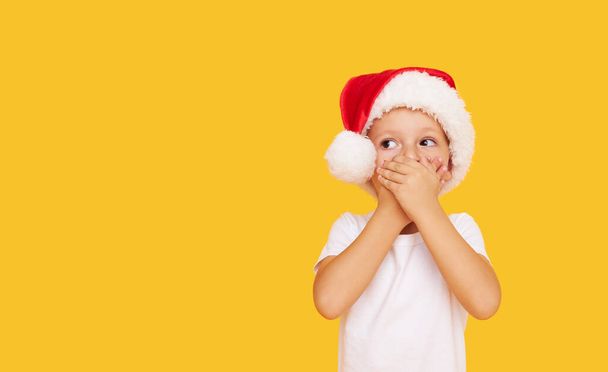A small child boy in a Santa Claus hat covers his mouth with his hands instead of a protective mask. Christmas and New Years holiday during the worldwide coronavirus pandemic - Foto, Imagem