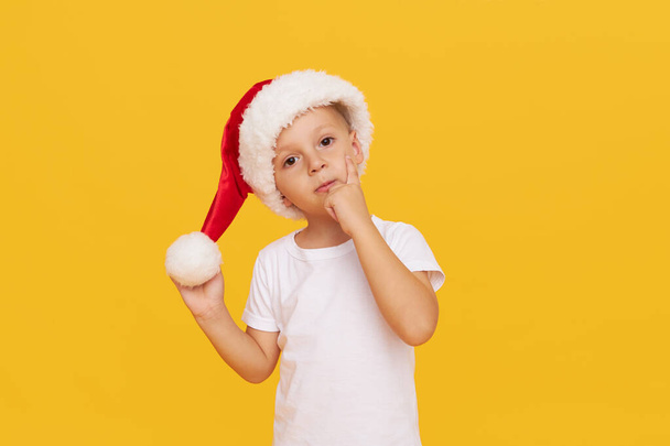 Portrait of Pensive little child boy dressed in christmas Santa hat. isolated over yellow background - Zdjęcie, obraz