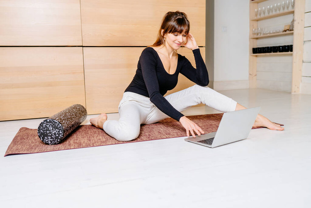 Gorgeous woman lying on mat and performing back exercise with foam roller at modern home interior. Female coach with laptop showing for patient fascia exercise on side surface of back. Online workout. - Foto, afbeelding