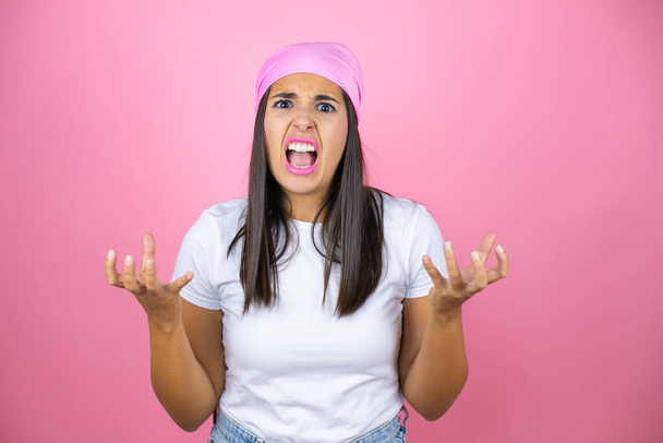 Jovem mulher bonita vestindo lenço de cabeça rosa sobre fundo rosa isolado louco e louco gritando e gritando com expressão agressiva e braços levantados. Conceito de frustração. - Foto, Imagem