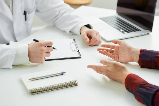 Μια κοντινή φωτογραφία των χεριών μιας γυναίκας ασθενούς και ενός γιατρού κατά τη διάρκεια ενός ραντεβού σε ένα νοσοκομείο. Ένα σημειωματάριο με στυλό, ένα ντοσιέ, ένα λάπτοπ βρίσκονται σε ένα λευκό γραφείο στο γραφείο ενός γιατρού.. - Φωτογραφία, εικόνα