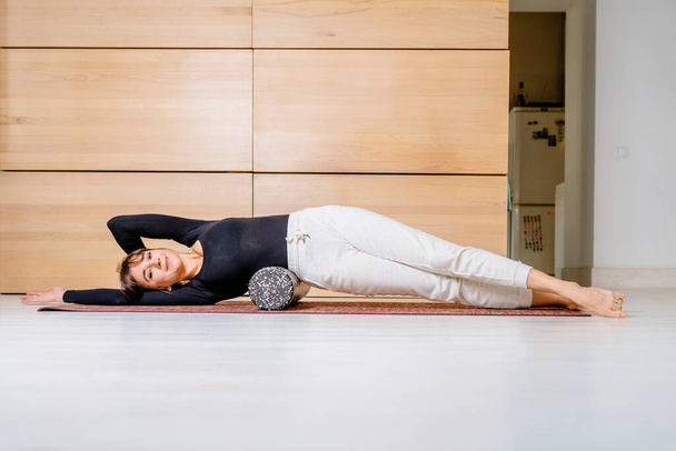 Brunette female yoga pilates instructor in black shirt white trousers costume doing exercise with foam roller for massage in modern cozy home interior. yoga and healthy lifestyle theme concept. - Foto, imagen