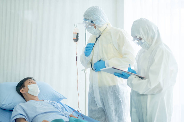 Γιατρός με ολόσωμη στολή κάλυψης εξηγεί την εξέλιξη της νόσου του ασθενούς και ο νοσηλευτής καταγράφει τα δεδομένα δίπλα στο κρεβάτι του ασθενή Covid- 19. - Φωτογραφία, εικόνα