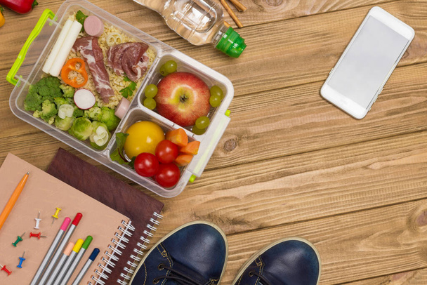 Blue shoes, notebooks with pens and bacon and vegetable lunchbox. Light wooden background. View from above. Copy space - Photo, image