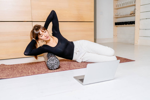 Fitness virtual online exercise concept. Woman instructor streaming from internet at home. Sporty girl in sportswear working out. Workout training in living room, laptop and foam roller a side. - Photo, Image