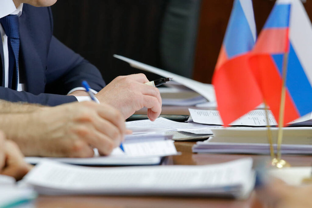 Un uomo in giacca e cravatta ha una penna stilografica. Sul tavolo ci sono documenti aziendali, cartelle, quaderni e la bandiera russa - Foto, immagini