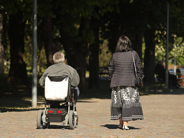 Eine Frau geht mit einem Mann, der mit einem elektrischen Rollstuhl unterwegs ist, im Park spazieren. Bedürftige in Obhut nehmen. Die Aktivität von Menschen mit eingeschränkter Mobilität. Prothetik von Patienten des Bewegungsapparates. - Foto, Bild