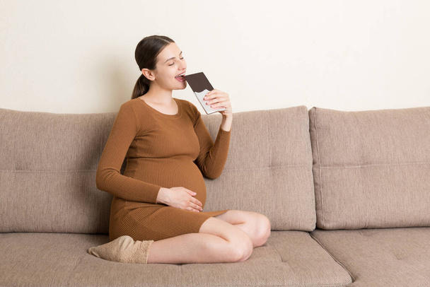 Una donna incinta che si rilassa sul divano sta mangiando una tavoletta di cioccolato a casa. Cibo malsano durante il concetto di gravidanza. - Foto, immagini