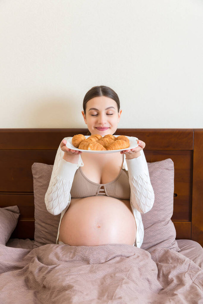 Genç hamile bir kadın, süt ve kruvasan kahvaltısı yapıyor, yatakta oturuyor ve karnına bakıyor, boşluğa. - Fotoğraf, Görsel