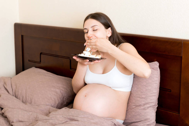 Hungry pregnant woman relaxing in bed is eating greedily a piece of cake and has a dirty mouth. Expecting mother can't stop eating. Strong appetite during pregnancy concept. - Photo, Image