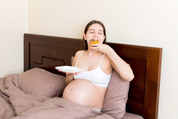 Mulher grávida com fome relaxando na cama está comendo gananciosamente um pedaço de bolo e tem uma boca suja. Esperar que a mãe não pare de comer. Forte apetite durante o conceito de gravidez. - Foto, Imagem