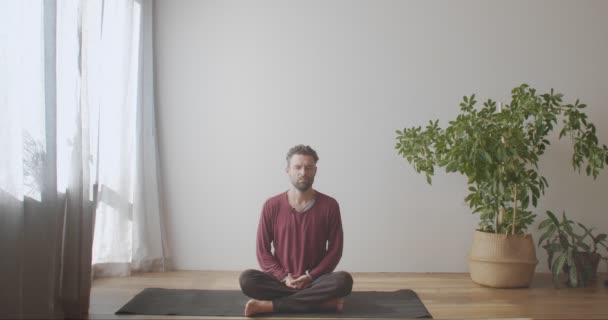 Hombre barbudo sentado con las piernas cruzadas en la esterilla de yoga practicando pranayama con los ojos cerrados. Joven maestro yogui caucásico haciendo ejercicios de respiración en interiores luz natural cámara lenta. hábitos de estilo de vida saludable - Metraje, vídeo