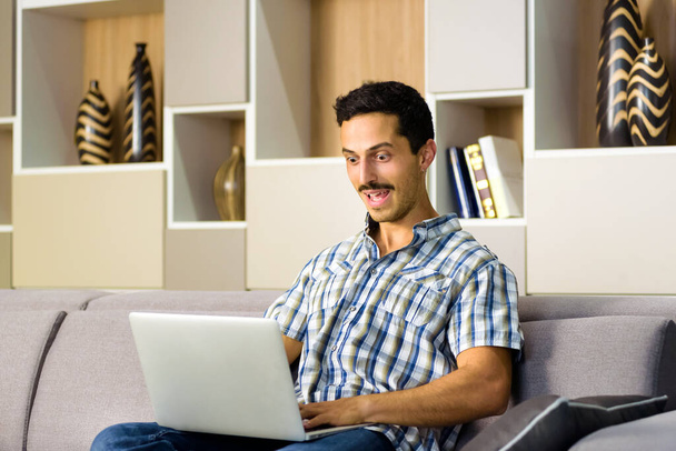 Nuori mies reagoi hämmästyneenä kannettavaan tietokoneeseen, joka tuijottaa leveäsilmäistä suutaan agape kun hän rentoutuu kotona mukavalla sohvalla - Valokuva, kuva