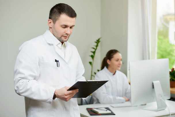 Un chirurgien avec des soies en manteau blanc tient un presse-papiers noir avec des documents et un stylo dans un hôpital. Une femme médecin assise devant l'ordinateur de bureau dans le coin de la pièce.  - Photo, image