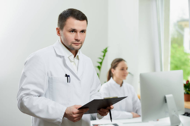 Серйозний хірург з щетиною в білому пальто тримає чорний кишеню з документами і ручкою в лікарні. Жінка-лікар сидить перед настільним комп'ютером у кутку кімнати
.  - Фото, зображення