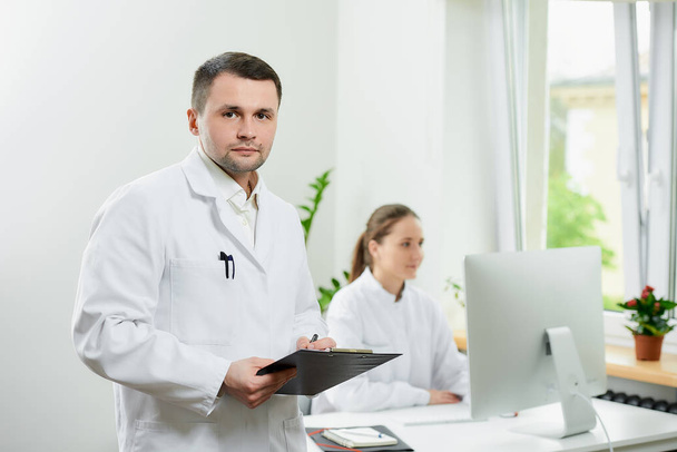 Vážný chirurg se štětinami v bílém plášti drží černou podložku s dokumenty, které zůstávají v nemocnici. Ženská doktorka sedící před stolním počítačem v rohu místnosti.  - Fotografie, Obrázek