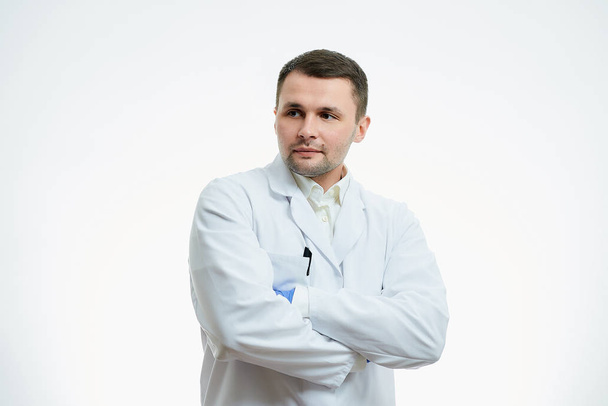 A male caucasian doctor in a white lab coat and blue disposable medical gloves is staying crossed his arms over his chest. A scientist is isolated over white background. - Fotoğraf, Görsel