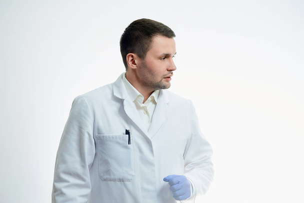 Un médico caucásico con una bata blanca de laboratorio y guantes médicos desechables azules giró la cabeza hacia un lado. Un científico está aislado sobre fondo blanco. - Foto, imagen