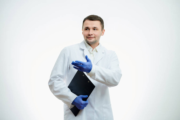 Un gentil médecin en blouse blanche et des gants médicaux jetables bleus tient un carton d'une main et l'autre pointe du doigt. Un scientifique est isolé sur fond blanc. - Photo, image