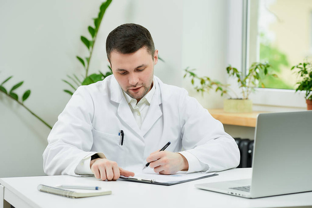 Ένας λευκός γιατρός με άσπρη ποδιά συμπληρώνει μια κάρτα ασθενούς με μια διάγνωση κοντά σε ένα λάπτοπ στο νοσοκομείο. Ένας θεραπευτής περιμένει έναν ασθενή στο γραφείο ενός γιατρού.. - Φωτογραφία, εικόνα