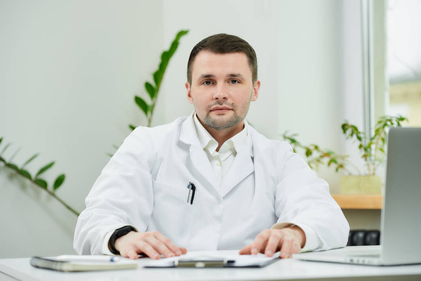 Egy kedves kaukázusi orvos fehér köpenyben az asztalánál pihen, és intenzíven hallgat a kórházban. Egy terapeuta vár egy betegre egy laptop mellett az orvosi rendelőben.. - Fotó, kép
