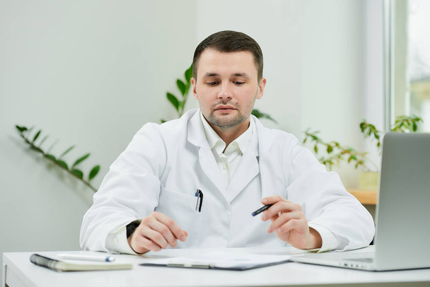 Un médico caucásico con una bata blanca de laboratorio está sentado en su escritorio y bajó los ojos durante el examen de la historia clínica del paciente en un hospital. Un terapeuta cerca de un portátil en el consultorio de un médico. - Foto, imagen