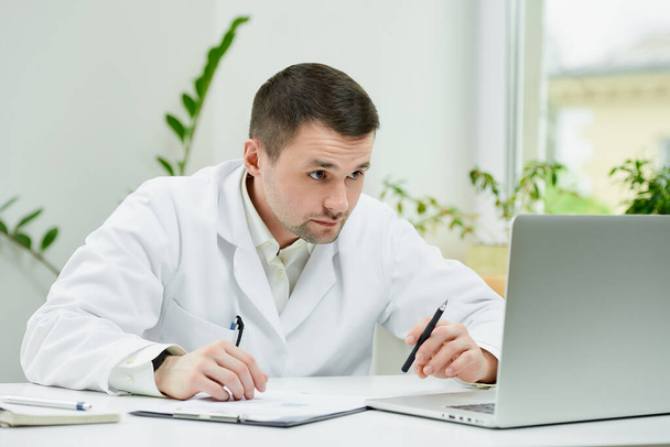Vážný běloch v bílém laboratorním plášti zapisuje pacientovu anamnézu do počítače v nemocnici. Terapeut sedí u stolu s laptopem v ordinaci lékaře.. - Fotografie, Obrázek